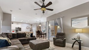 A modern living room with a sectional sofa, ottoman, and a glass coffee table. There is a dining area with a table and chairs, and a kitchen in the background. - Experience magic! HR Golf Course view 5bd/3b w/pool-spa near all Disney Parks (Davenport)