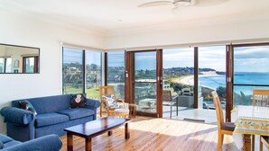 A spacious living room with a large sliding glass door leading to a balcony with a view of the beach and ocean. - 18 Cliff Road - Opposite One Mile Beach! (Forster)
