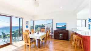 A spacious dining area with a large table and chairs, a television, and a view of the sea through large windows. - 18 Cliff Road - Opposite One Mile Beach! (Forster)