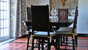 A dining area with a table set for four, a stone wall with a cross, and a window with patterned curtains.