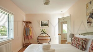 Un dormitorio con cama, un radiador, una ventana con persianas, un estante de madera con toallas, un cuadro en la pared y una puerta que da a otra habitación.