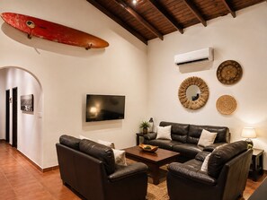 A living room with a leather sofa set, a flat-screen TV, and a surfboard decoration. - Villa Colibrí, 3 BR, walk to beach, private pool (Santa Teresa Beach)