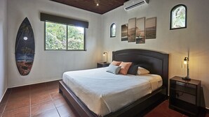 A bedroom with a bed, a nightstand, a window, and a surfboard decoration. - Villa Colibrí, 3 BR, walk to beach, private pool (Santa Teresa Beach)