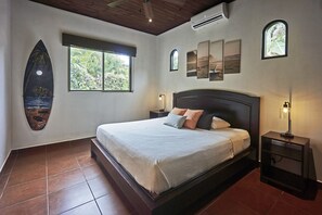 A bedroom with a bed, a nightstand, a window, and a surfboard decoration. - Villa Colibrí, 3 BR, walk to beach, private pool (Santa Teresa Beach)