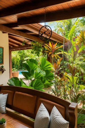 Teto de madeira com luminária pendente e vista para um jardim exuberante, com diversas plantas e uma piscina.