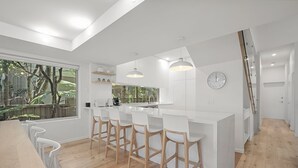 A modern kitchen with a large island, bar stools, a clock, and a view of the outdoors. - Audrey - Little Cove (Noosa Heads)