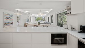 A modern kitchen with white cabinetry, a central island, built-in appliances, pendant lighting, and large windows. - Audrey - Little Cove (Noosa Heads)