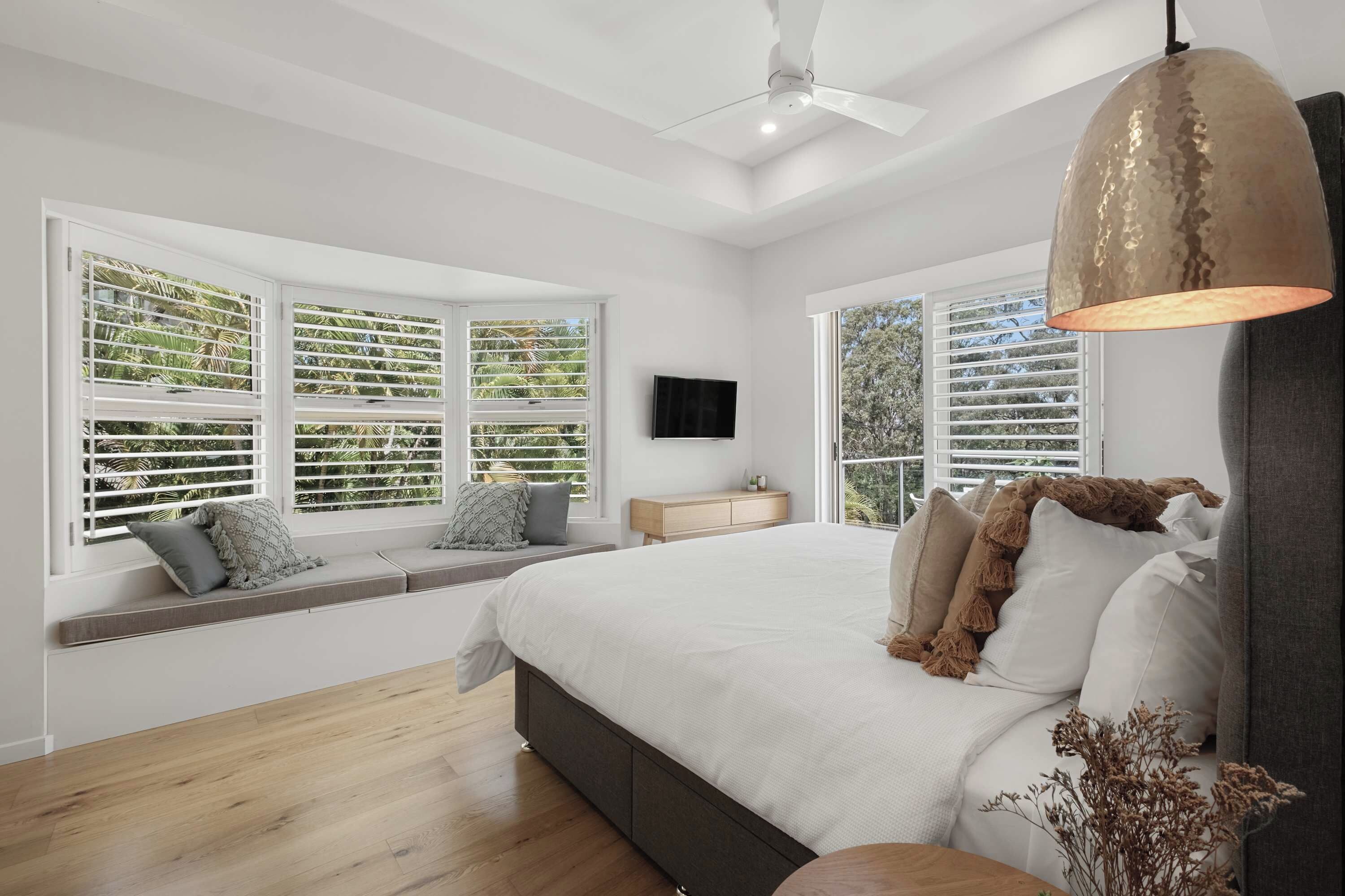 A bedroom with a large bed, a ceiling fan, and a view of greenery through the windows.