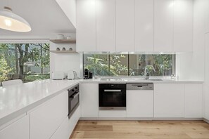 A modern kitchen with white cabinetry, a built-in dishwasher, and a large window overlooking a garden. - Audrey - Little Cove (Noosa Heads)