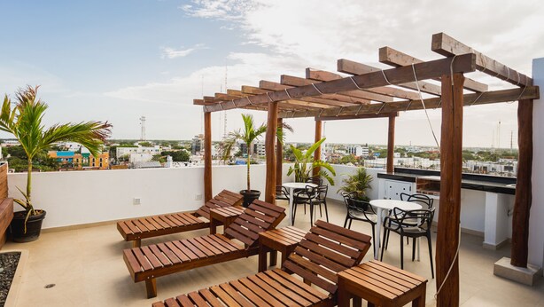 Una terraza en la azotea con tumbonas de madera, una pérgola y una pequeña zona de comedor con sillas y mesas.