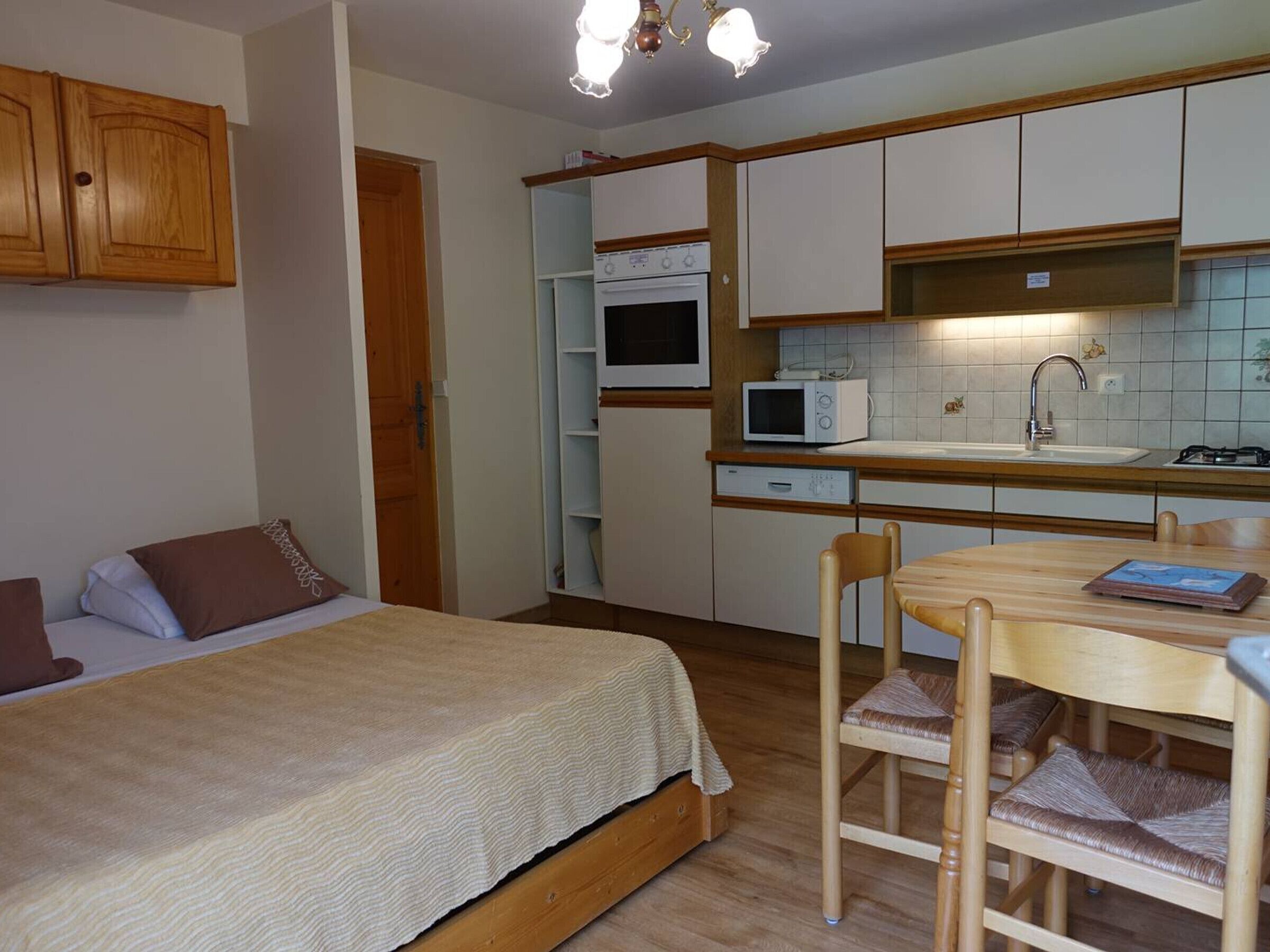 A compact kitchen with a microwave, stove, and sink. There is a bed with pillows and a wooden table with chairs.