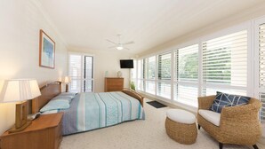 A bedroom with a bed, bedside tables, a TV, a wicker chair, and large windows with blinds. - Le Point 101 - Walking distance to Main Beach (Forster)