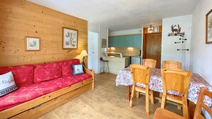 Una sala de estar acogedora con un sofá rojo, una mesa de comedor de madera y sillas. Se ve una cocina al fondo con armarios color turquesa.