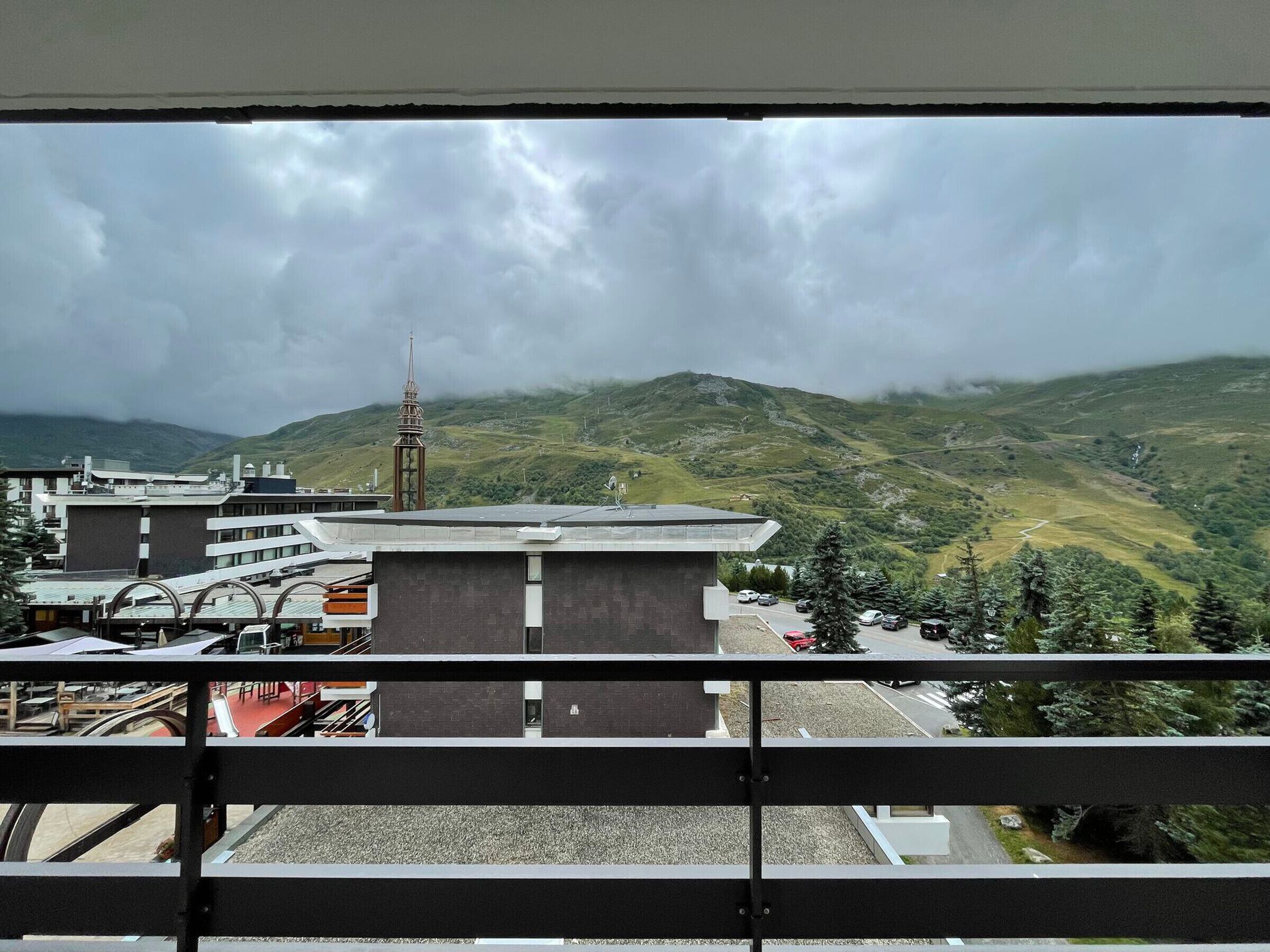 A view from a balcony overlooking a mountainous landscape with a church spire, modern buildings, and a parking area.