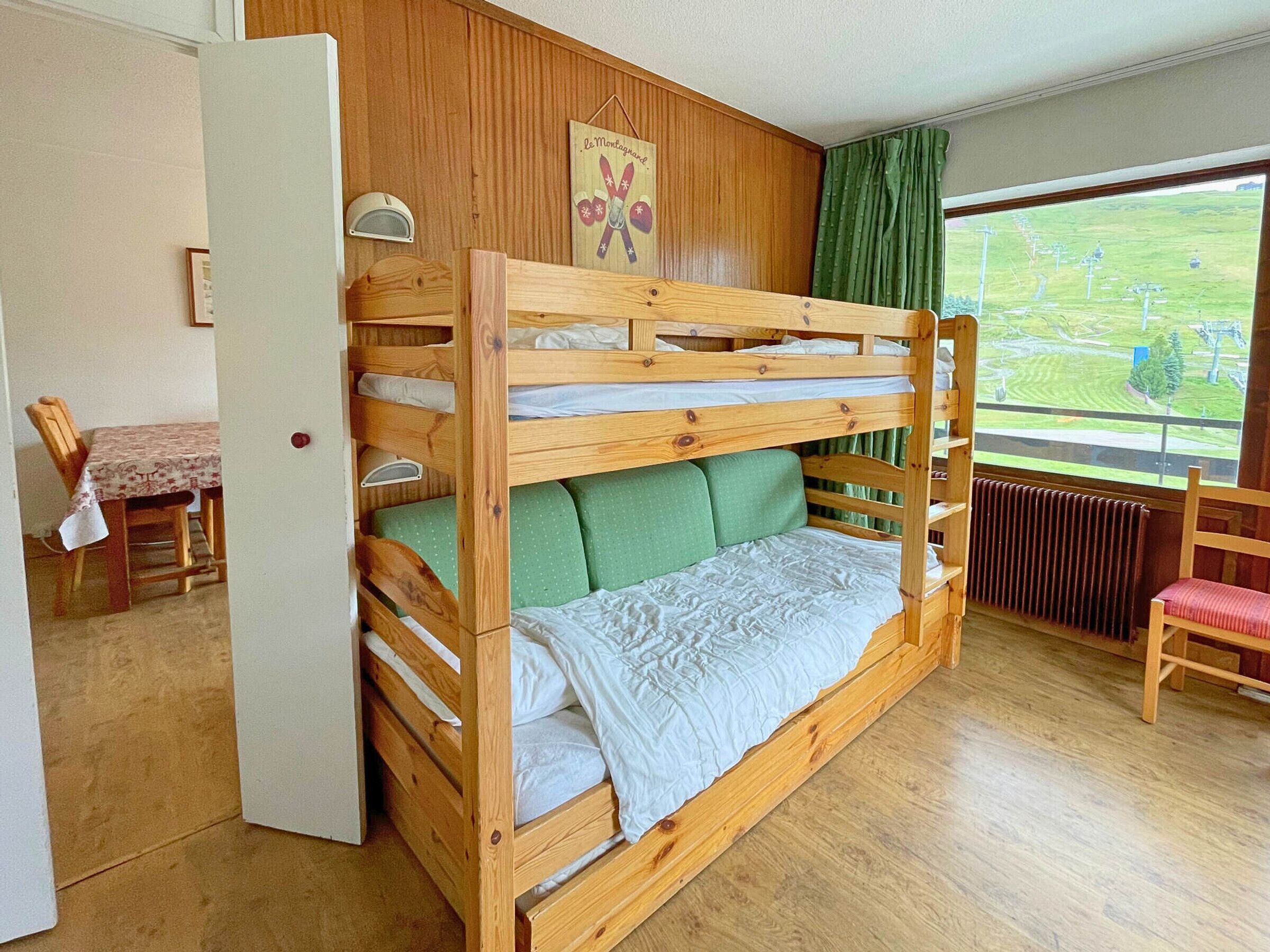 A bunk bed with a green cushion, a wooden table with a floral tablecloth, and a view of a green field through a window.
