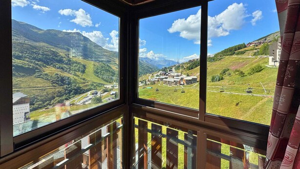 A room with large windows offering a view of a mountainous landscape and a small village. - Cozy 3-Room Apartment for 5 with Ski-In/Ski-Out, Balcony, and Modern Amenities (Les Menuires)
