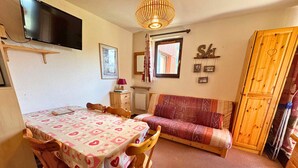 A cozy room with a dining table, chairs, a sofa, a TV, and a wooden cabinet. - Cozy 3-Room Apartment for 5 with Ski-In/Ski-Out, Balcony, and Modern Amenities (Les Menuires)