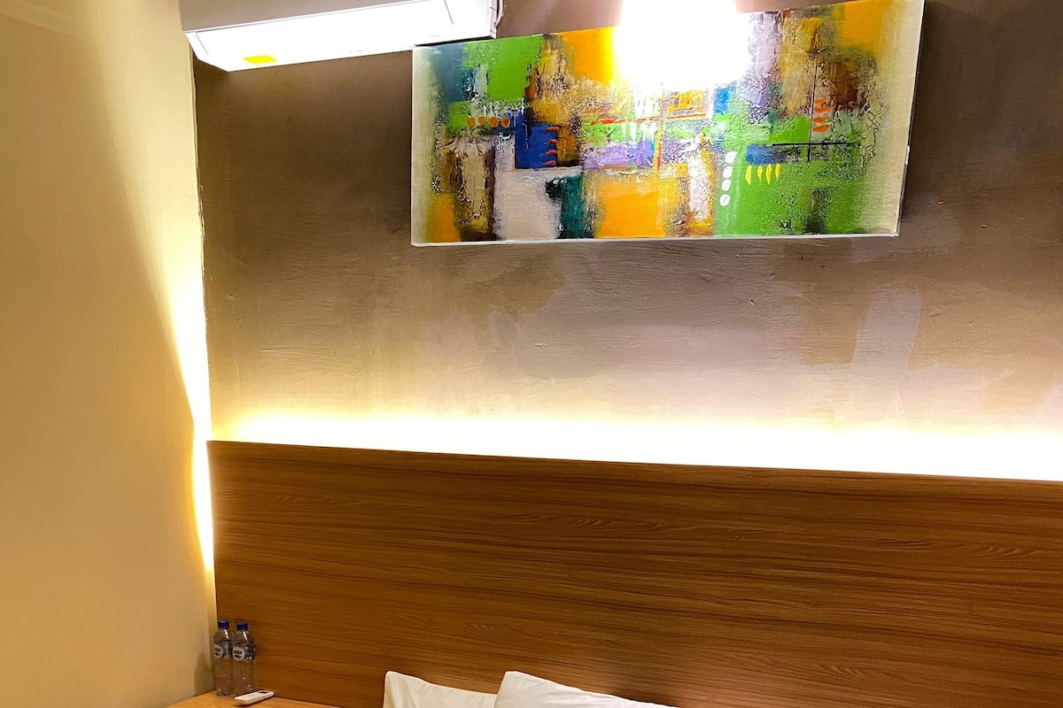 A hotel room with a neatly made bed, a wooden headboard, a nightstand with a lamp, and a painting on the wall.