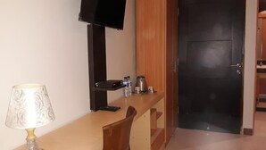 Een hotelkamer met een houten bureau, een aan de muur bevestigde televisie, een lamp, een koffiezetapparaat en een deur.