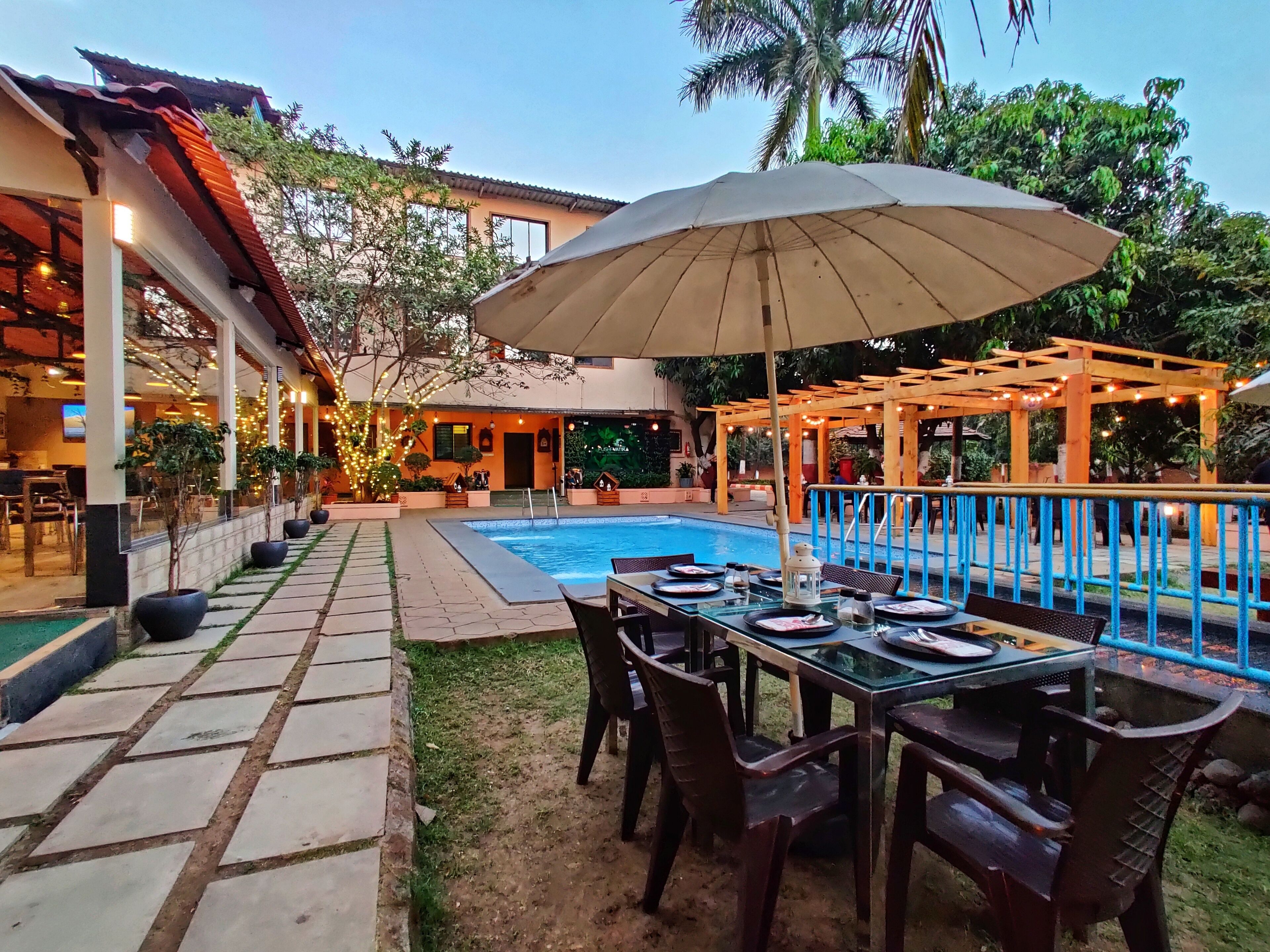 Un espace aménagé au bord de la piscine, avec une table dressée pour les repas, entourée de chaises, et une pergola décorée de guirlandes lumineuses.