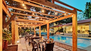 Un patio exterior con mesas y sillas, una piscina y una pérgola de madera con luces colgantes.