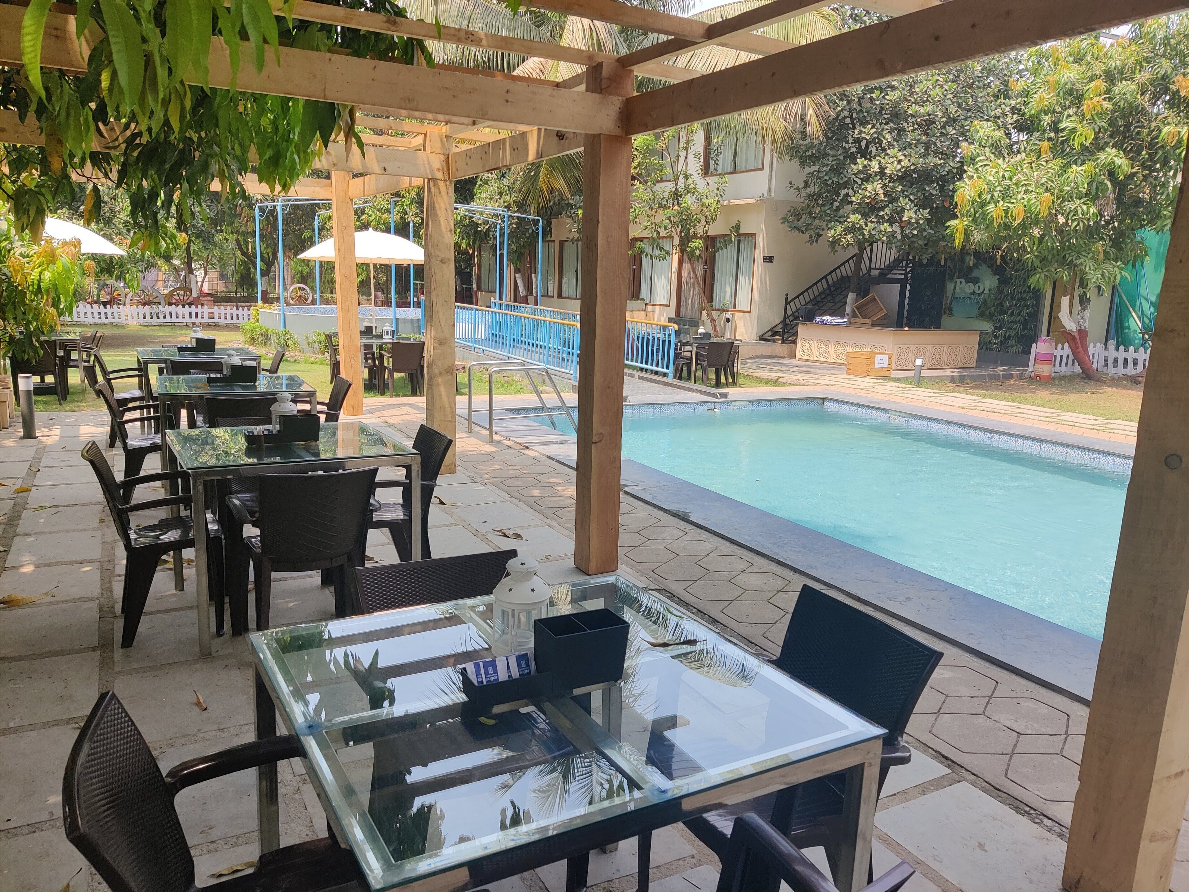A covered outdoor seating area with glass tables and chairs, a pool, and a view of a building with a blue railing.