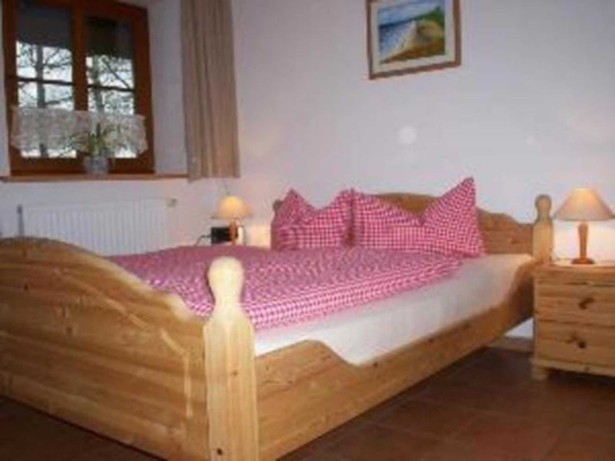 Uma cama de madeira com colcha e almofadas rosa xadrez, uma mesa de cabeceira com abajur e uma janela com vista para árvores.