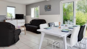 Un salon moderne comprenant un coin repas, un canapé en cuir noir, une table à manger blanche pour quatre personnes et un téléviseur posé sur un meuble.