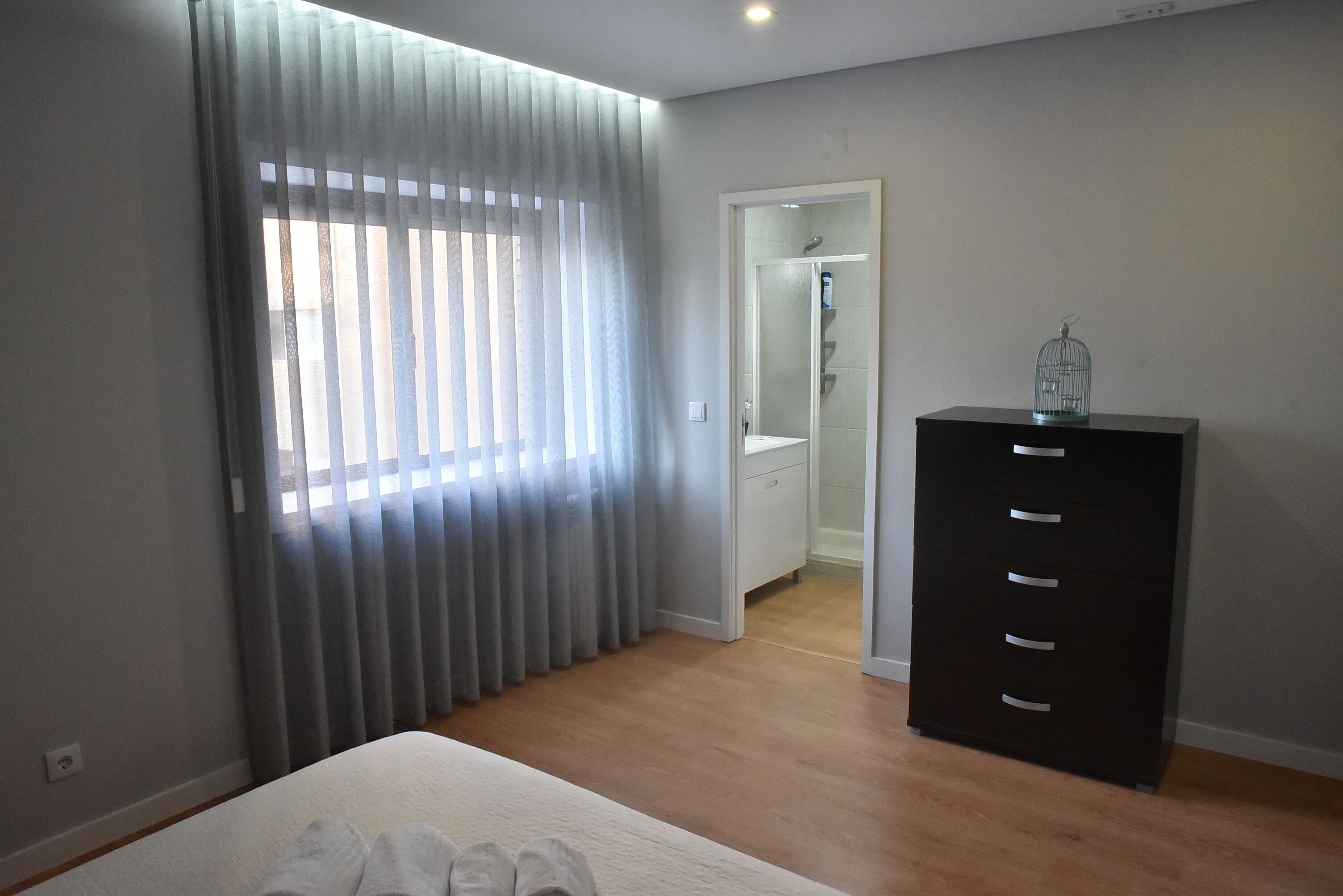 A bedroom with a bed, a dresser, a window with curtains, and a door leading to a bathroom.