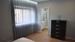 Un dormitorio con cama, cómoda, ventana con cortinas y puerta que da a un baño.