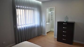 A bedroom with a bed, a dresser, a window with curtains, and a door leading to a bathroom.