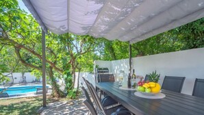 Un área de comedor exterior con piscina, cubierta por una marquesina blanca y rodeada de follaje.