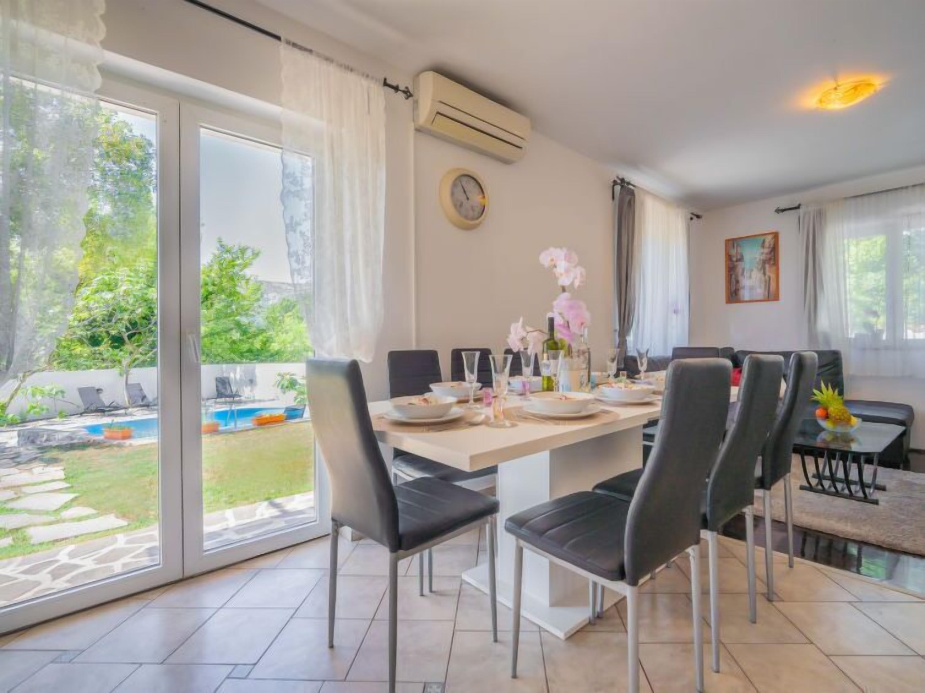 A dining area with a large table, chairs, and a view of a swimming pool and garden.