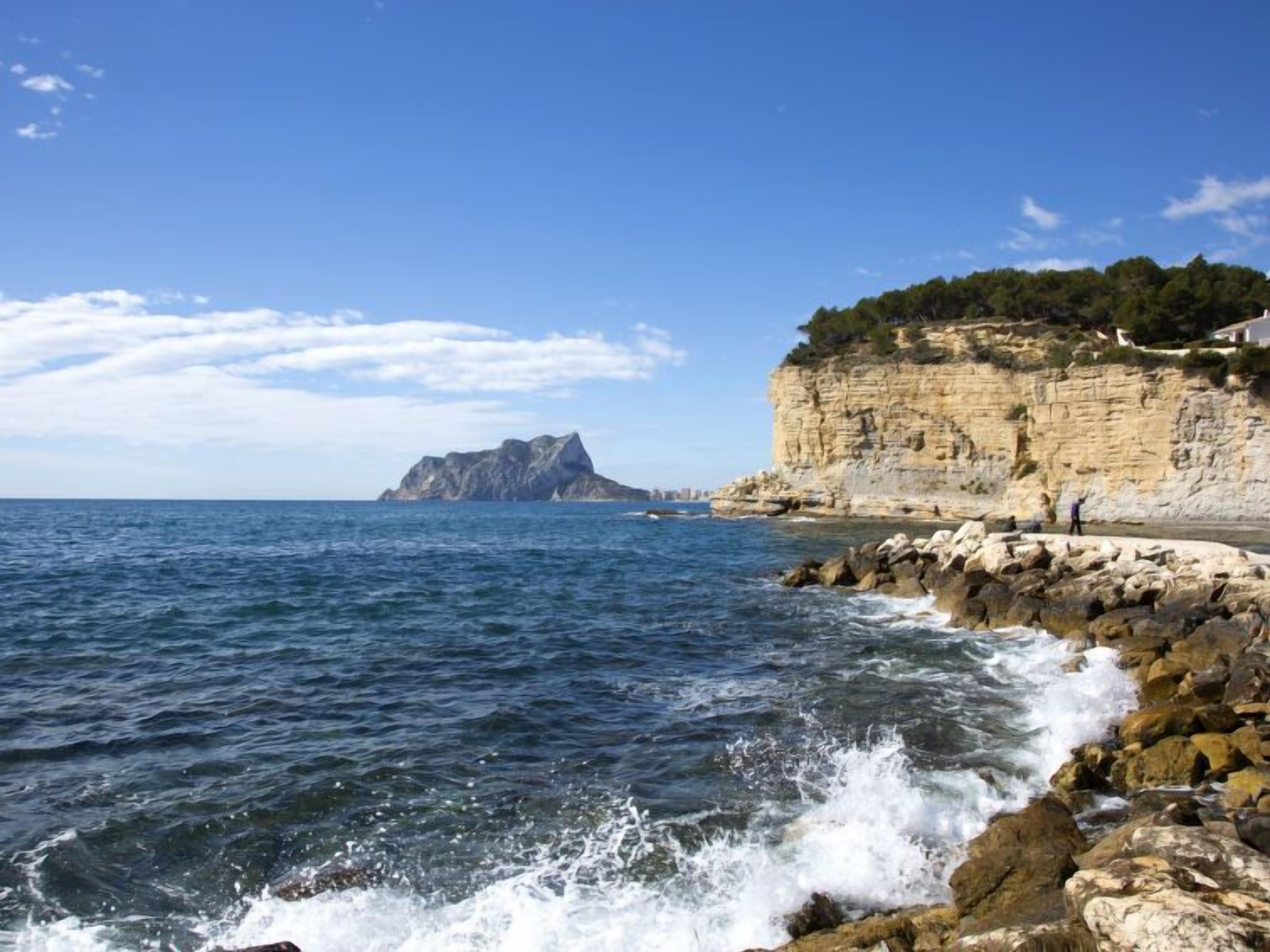 A coastal scene with a rocky shoreline, clear blue sea, and a prominent cliff with vegetation.