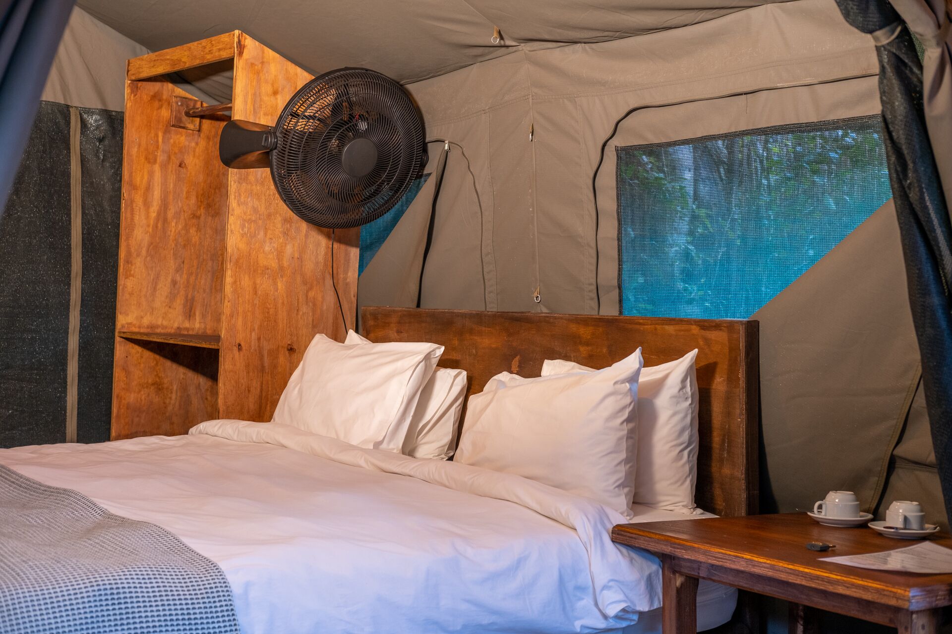 A tented room with a wooden bed, white bedding, and a table with a cup and saucer.