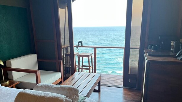 A room with a bed, a wooden bench, a chair, and a view of the ocean. - Sky Island Resort (Ponta d'Ouro)
