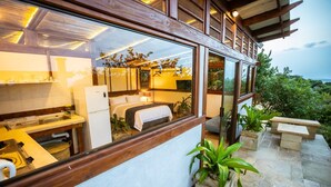 A modern hotel room with a large window, a bed, a small kitchen area, and a stone patio with plants. - Sky Island Resort (Ponta d'Ouro)