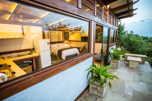 Una habitación de hotel moderna con un ventanal grande, una cama, una kitchenette y un patio de piedra con plantas.