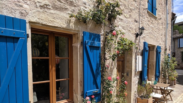 Un edificio de piedra con contraventanas azules, un banco de madera y una mesa con sillas en el exterior.