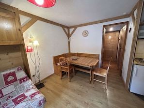 Ein kleiner Raum mit einem Holztisch, Bänken, einer kleinen Lampe, einer Wanduhr und einer Tür zu einem anderen Raum.