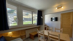 A room with a bed, a desk, and a chair. There is a window with curtains, a radiator, and a TV on the wall. - Classic apartment for 4 at La Croisette (Les Menuires)