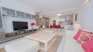 A modern living room with a flat-screen TV, a beige coffee table, and a white sofa with red pillows. There is a dining area with red chairs and a kitchen in the background.