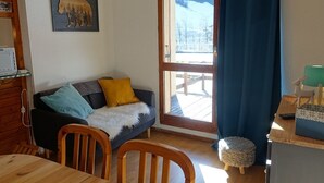 Ein gemütlicher Raum mit einem Holztisch, einer Couch mit einem Fellüberwurf, einem kleinen runden Hocker und einem Fenster mit Blick auf eine verschneite Landschaft.