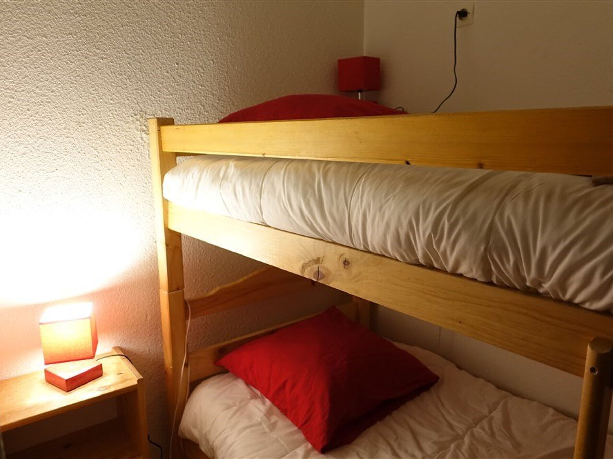 Una cama litera con un almohadón rojo y una mesita de noche con una lámpara.