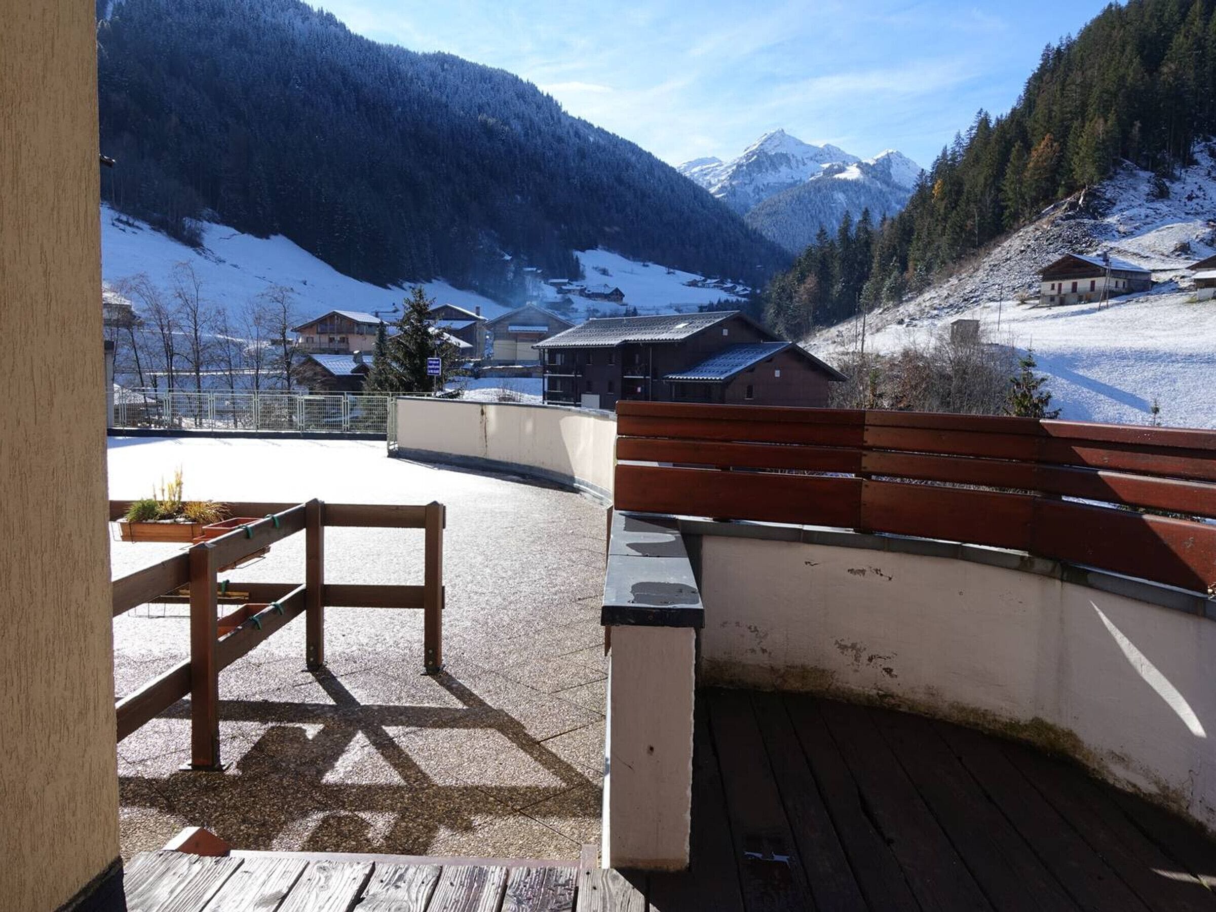 Una terraza de madera con un banco, una mesa y vistas a montañas nevadas y casas.