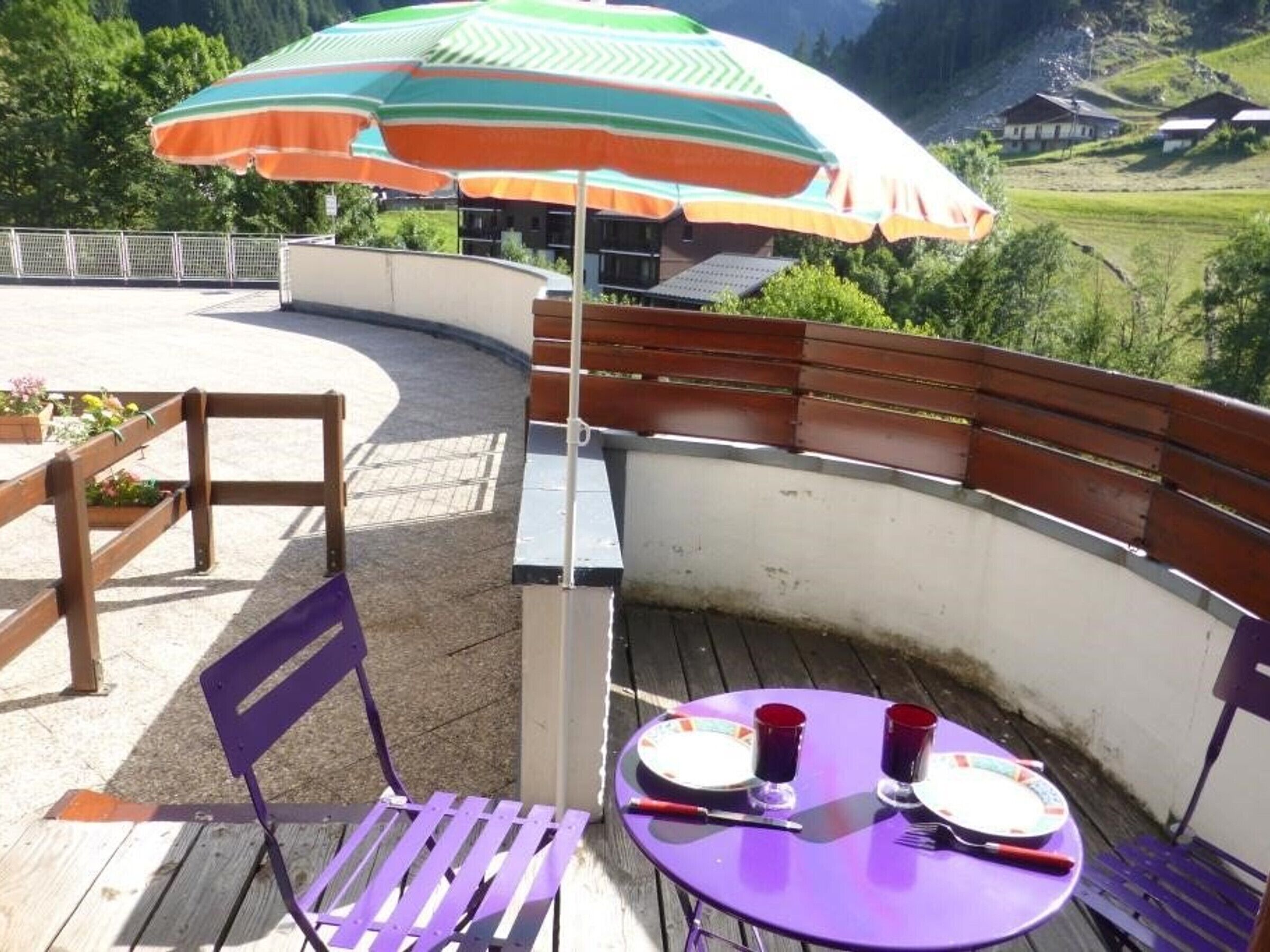 Eine Terrasse mit einem lila Tisch und Stühlen, einem Sonnenschirm und Blick auf eine gebirgige Landschaft.