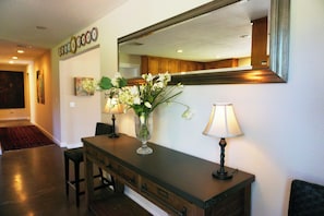 A reception area with a wooden console table, a vase of flowers, two wall-mounted lamps, a large mirror, and a red carpet leading to a door.