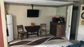 Un espace de vie compact comprenant un téléviseur fixé au mur, un réfrigérateur, un meuble en bois contenant des appareils de cuisine, deux chaises, un tapis à motifs et un canapé bleu.