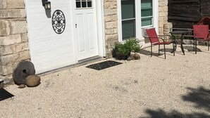 Un bâtiment en pierre avec une porte blanche, un petit porche avec des chaises rouges et une structure en bois en arrière-plan.