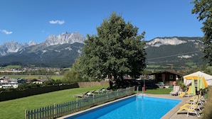 A swimming pool with a view of mountains, a large tree, and a wooden cabin. - Apartment Tirol 75m² with 2BR, Balcony, Pool & Mountain View - near Skislope (St. Johann in Tirol)